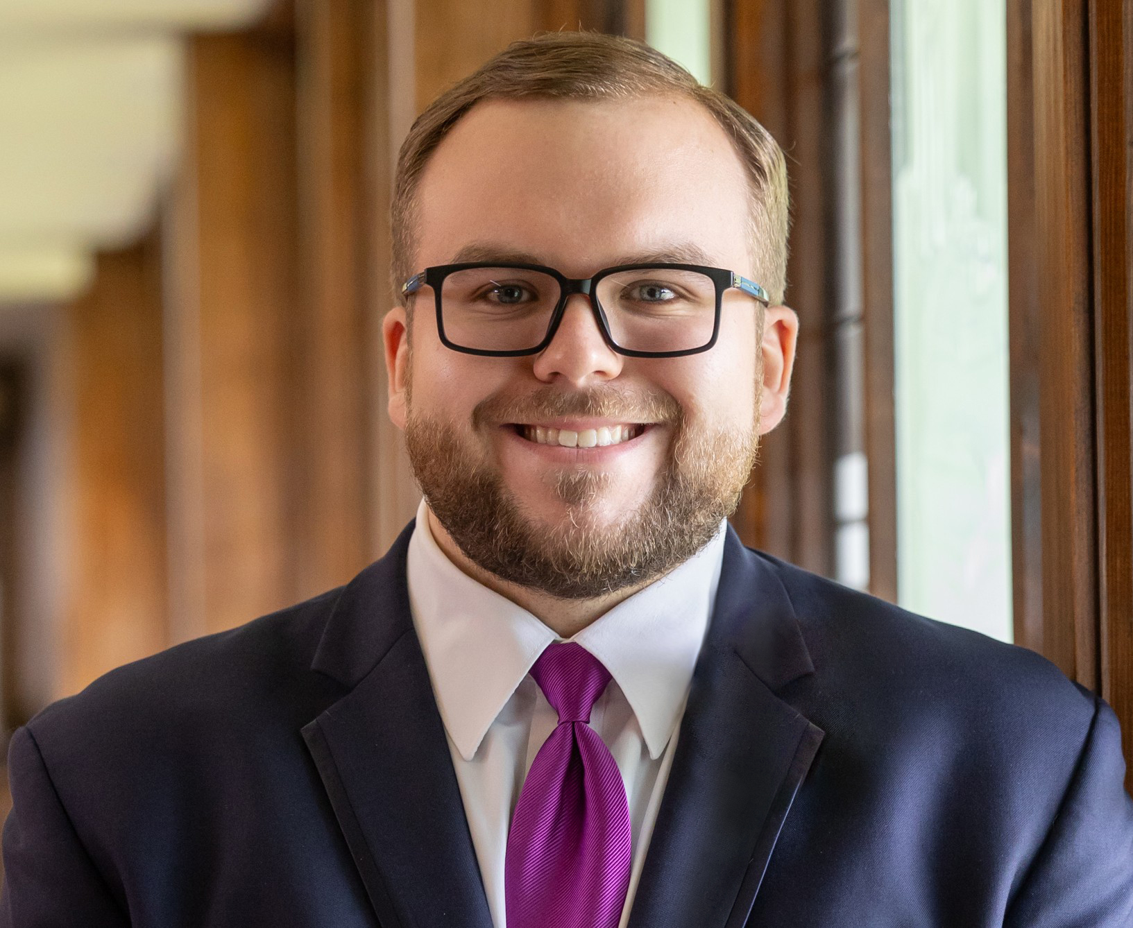 A photo of Chase Mieras. He has brown hair and blue eyes.
He is wearing a dark blue suit with a purple tie. He has a well trimmed beard, and wears glasses